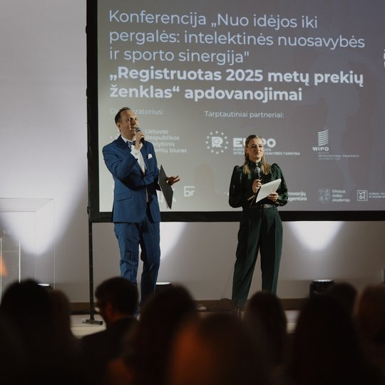 Nuotraukoje konferencijos salė su klausytojais. Tolumoje matomi renginio vedėjai ir  didelis ekranas.