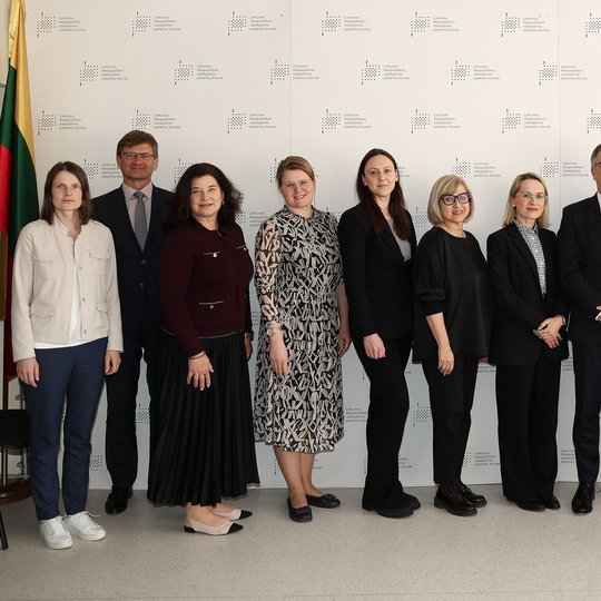 Nuotraukoje 11 susitikimo dalyvių. 3 vyrai ir 8 moterys. Už nugarų didelis stendas su Valstybinio patentų biuro besikartojančiu ženklu. Kairiame kampe stovi Lietuvos vėliava.